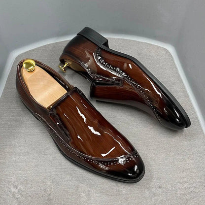 Glossy brown slip-on men's dress shoes with brogue detailing on a light fabric surface