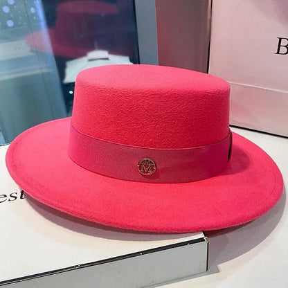 Bright pink flat top fedora hat with ribbon and gold emblem on a display box