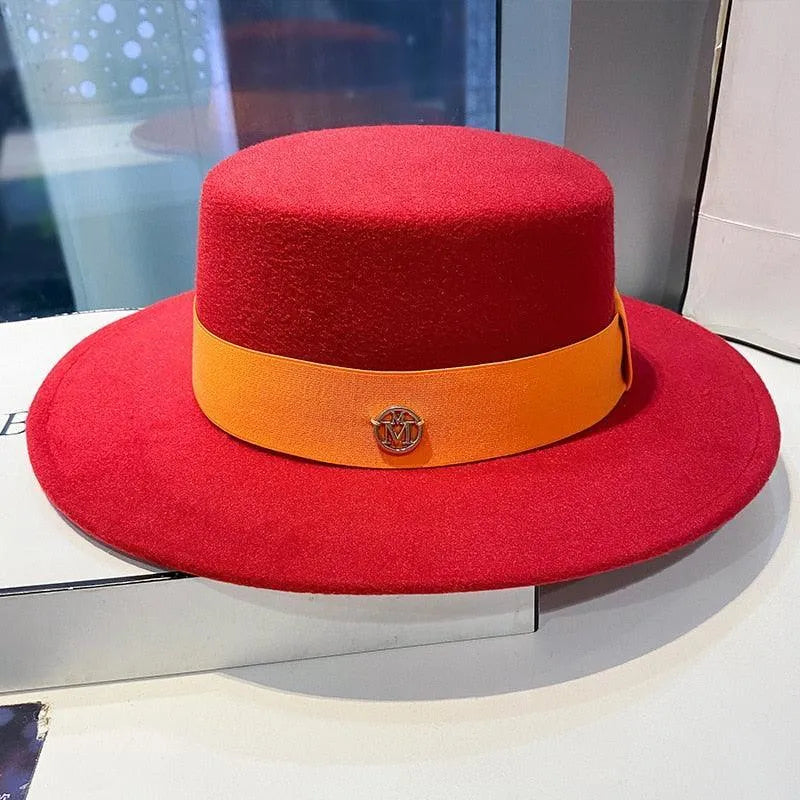 Red flat top fedora hat for women with orange band and gold emblem, displayed indoors